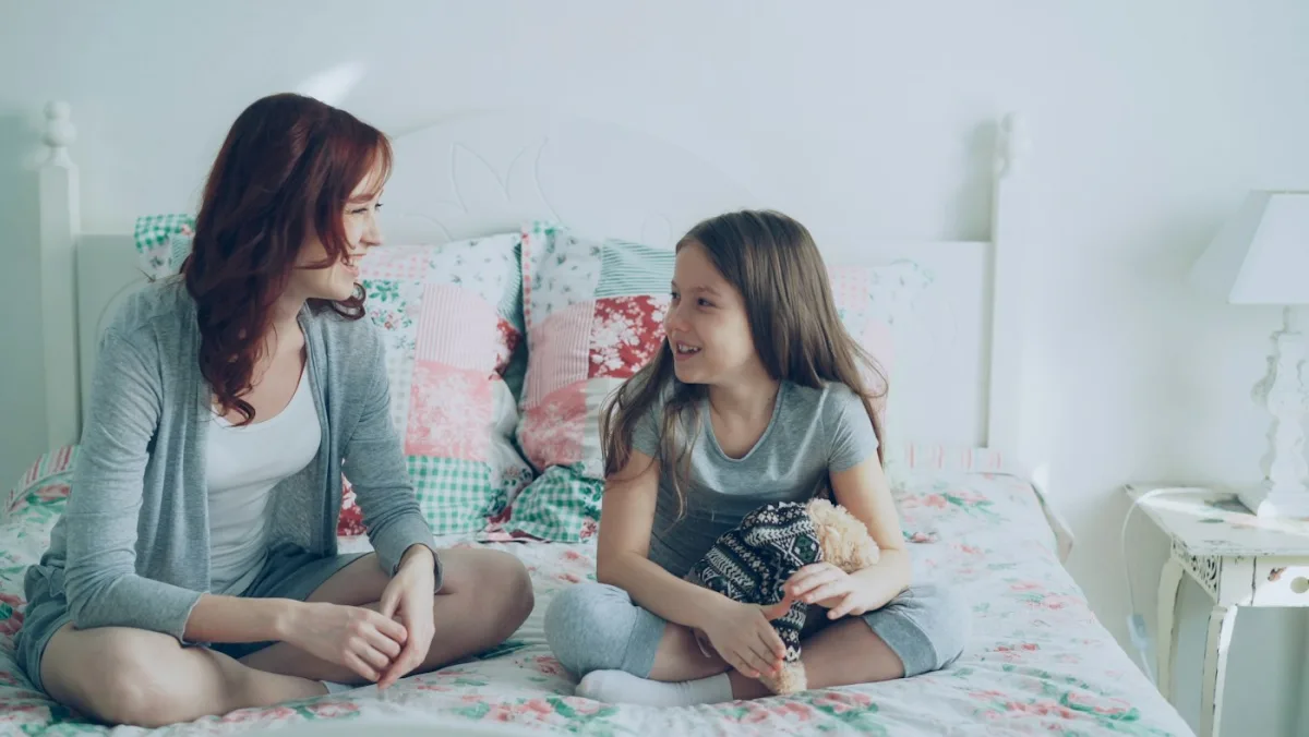 A parent and child sitting together on a bed, mid-conversation and smiling